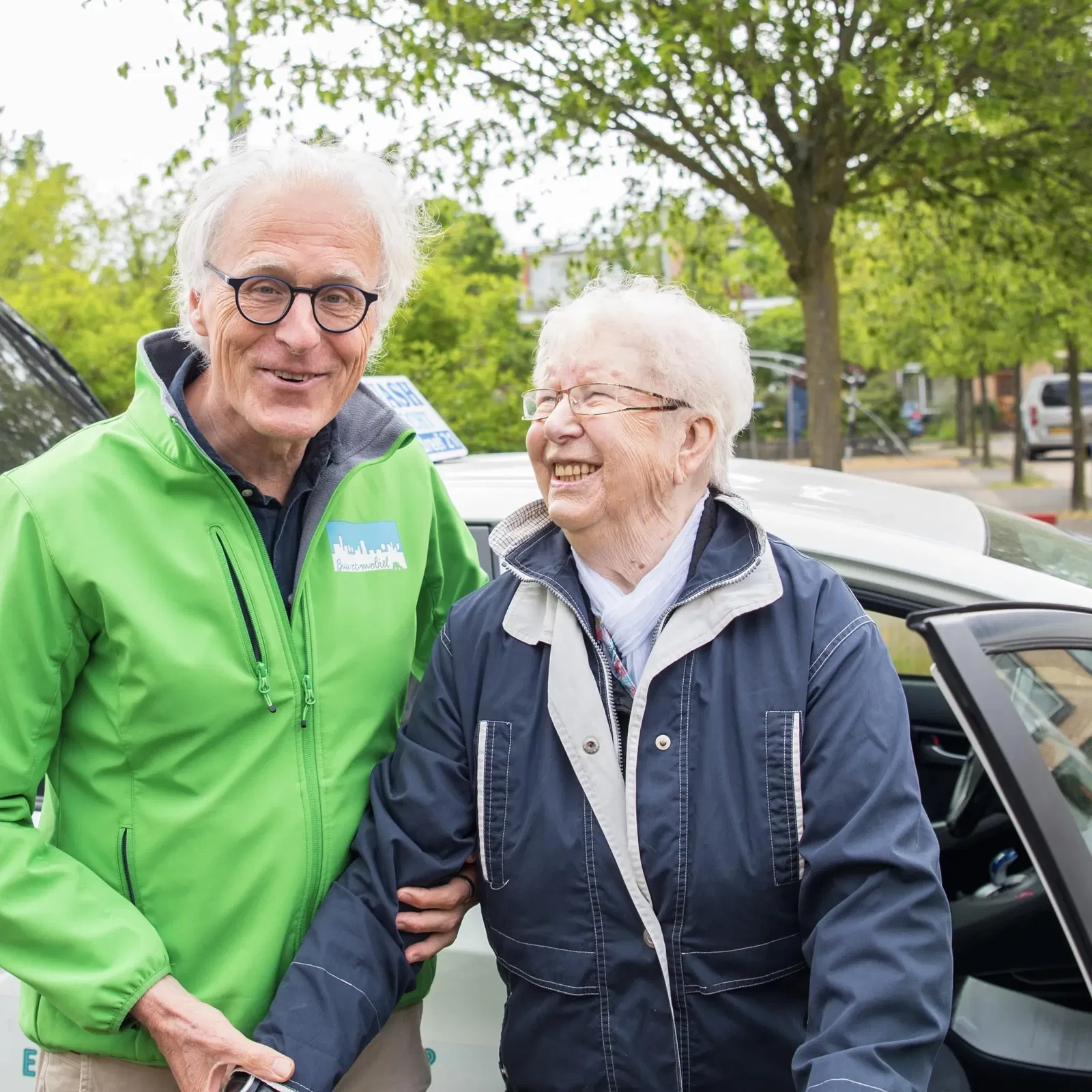 Een oudere vrouw die lacht en naast een chauffeur staat van Buurt Mobiel. De chauffeur draagt een groene jas met een logo van "Buurt Mobiel". Beiden lijken vrolijk en staan voor een auto van Buurt Mobiel, een gemeenschapsvervoerservice. Er is een rustige straat op de achtergrond met bomen en geparkeerde auto's.