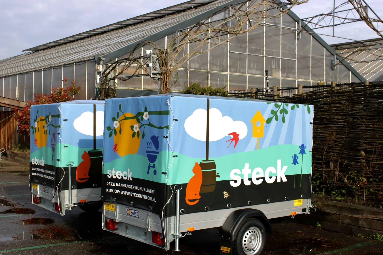 Aanhangwagen van tuincentrum Steck staat op het parkeerterrein voor de kas. De aanhangwagen heeft een kleurrijke en zomers gedecoreerde buitenkant, waar ook het logo van Steck op staat