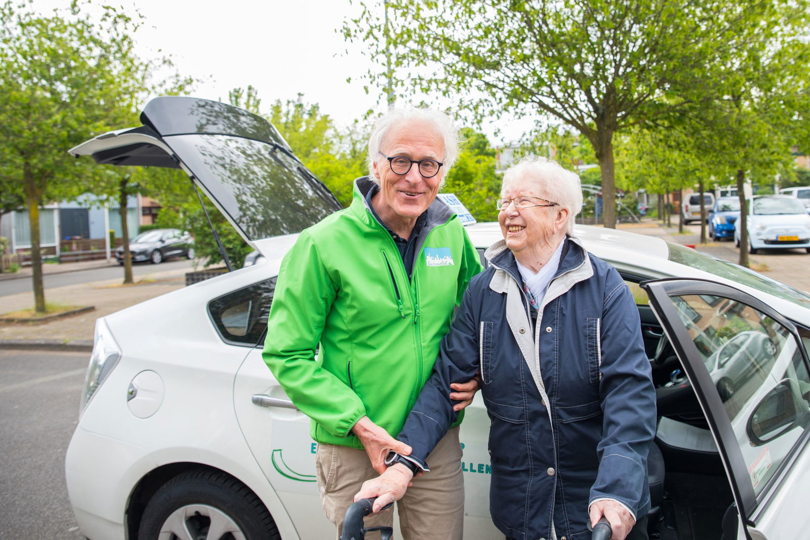 Een oudere vrouw die lacht en naast een chauffeur staat van Buurt Mobiel. De chauffeur draagt een groene jas met een logo van Buurt Mobiel. Beiden lijken vrolijk en staan voor een auto van Buurt Mobiel, een gemeenschapsvervoerservice. Er is een rustige straat op de achtergrond met bomen en geparkeerde autos.