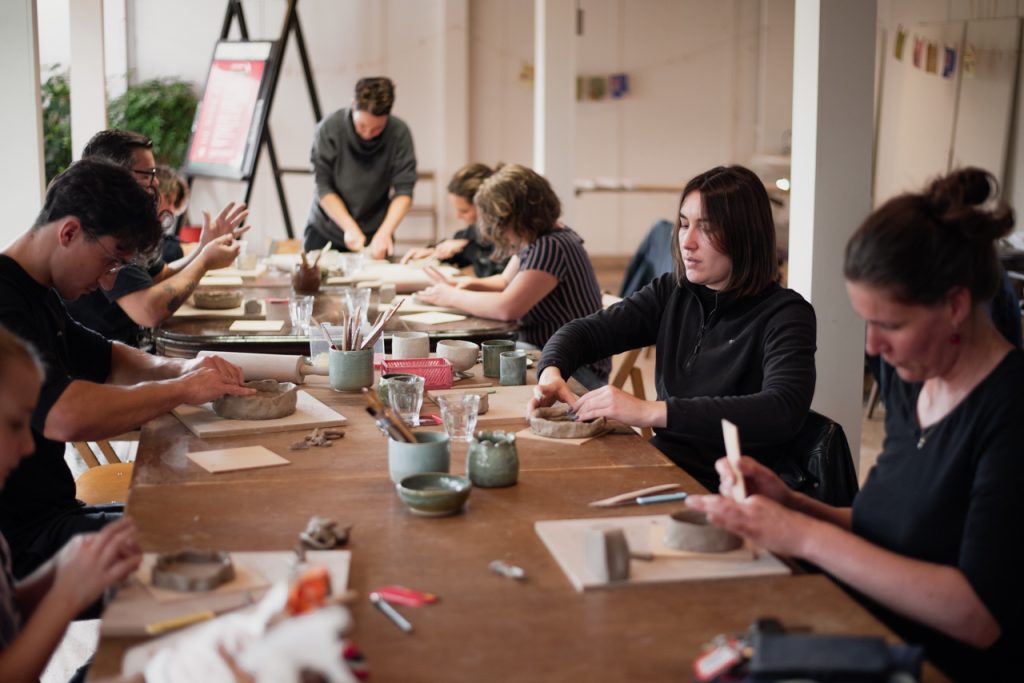 Een groep mensen zit aan een lange houten tafel en werkt geconcentreerd met klei. Ze zijn bezig met het vormen van verschillende objecten, omringd door gereedschap en potten met kleigereedschap. De sfeer is creatief en gefocust, met een gemengde groep van mannen en vrouwen van verschillende leeftijden die genieten van de workshop.