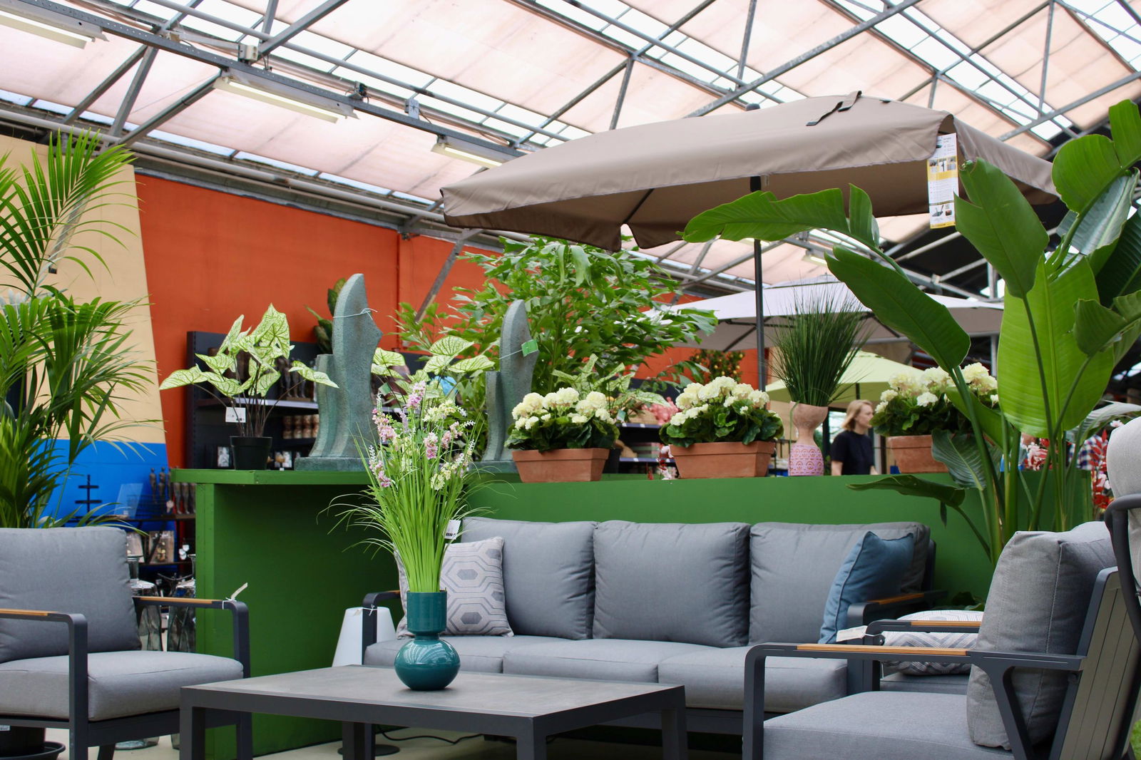 Een gezellige opstelling van tuinmeubels bij Steck in Utrecht Overvecht. De afbeelding toont een zithoek met comfortabele grijze kussens en een bijpassende tafel. Om de zithoek heen staan verschillende groene planten en bloemen in potten, die een frisse en levendige uitstraling geven. Boven de zithoek hangt een grote parasol die zorgt voor schaduw. De achtergrond laat een kleurrijke muur en het glazen dak van de kas zien. Op de achtergrond zijn klanten te zien die door de winkel lopen.