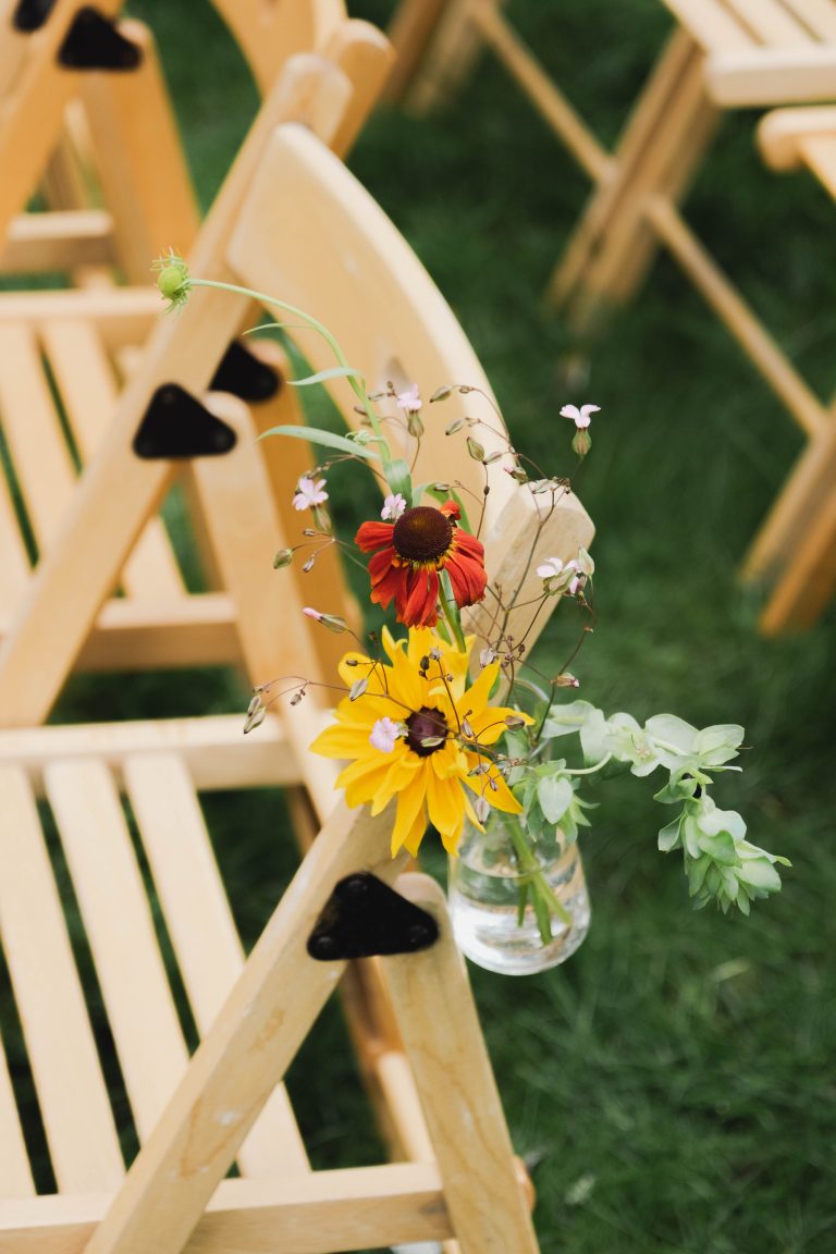 Een klein vaasje met een rood-oranje en een gele bloem uit Pluktuin Glitter Gladiool, vastgemaakt aan een houten stoel als decoratie voor de trouwceremonie. De styling is verzorgd door Noraly en geeft de setting een speelse en natuurlijke uitstraling.