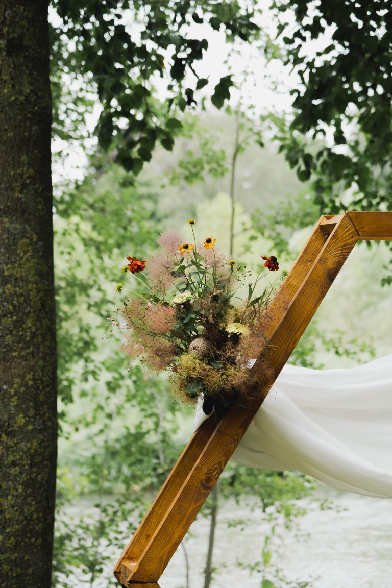 Een bloemstuk met wilde bloemen en pluizige takken van Pluktuin Glitter Gladiool, gestyled door Noraly. Het is bevestigd aan een zeshoekige houten trouwboog met een witte doek, geplaatst in een bosrijke omgeving.