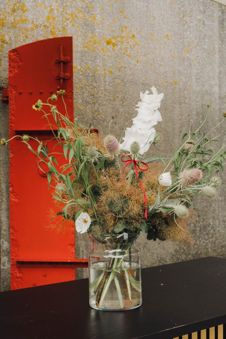Een natuurlijk bloemstuk met witte gladiool, distels en fijne takken, afkomstig van Pluktuin Glitter Gladiool en gestyled door Noraly. De vaas staat op een zwarte tafel voor een verweerde betonnen muur met een felrode deur.