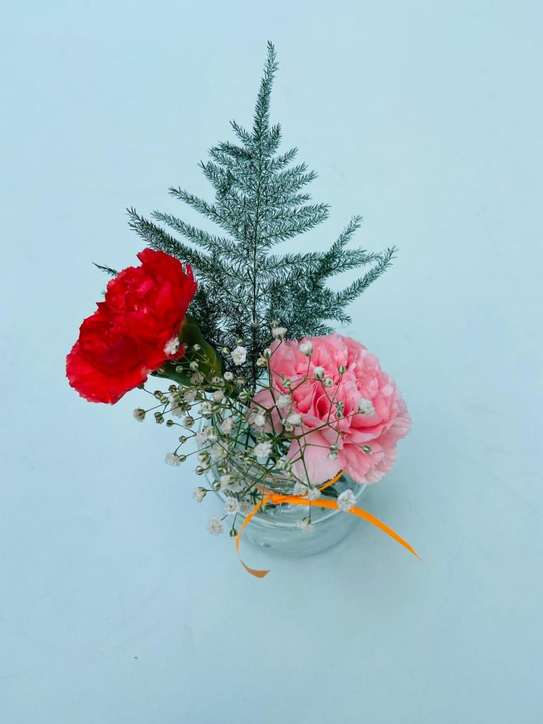 Een eenvoudig en elegant bloemstuk van Oogst met rode en roze anjers, gipskruid en groen blad, versierd met een oranje lint, in een glazen vaasje tegen een lichtblauwe achtergrond.