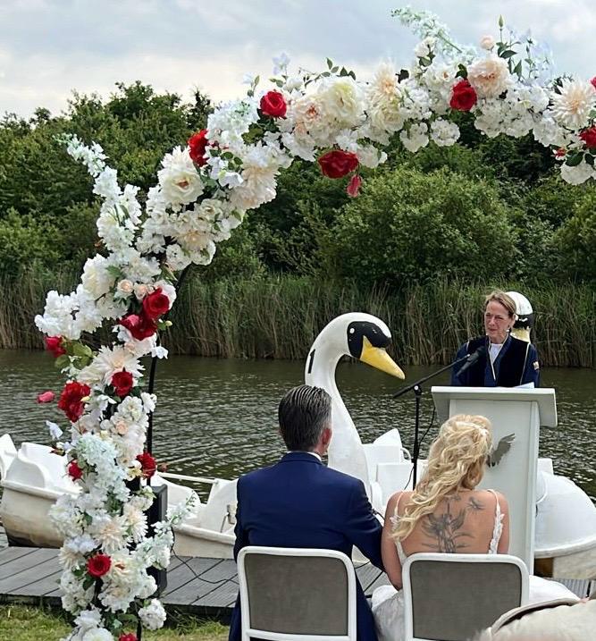 Een witte en rode bloemenboog gemaakt door oogst bij een trouwceremonie aan het water. Een bruidspaar zit voor een ceremonieel podium, met een groot zwanenbeeld in de achtergrond.