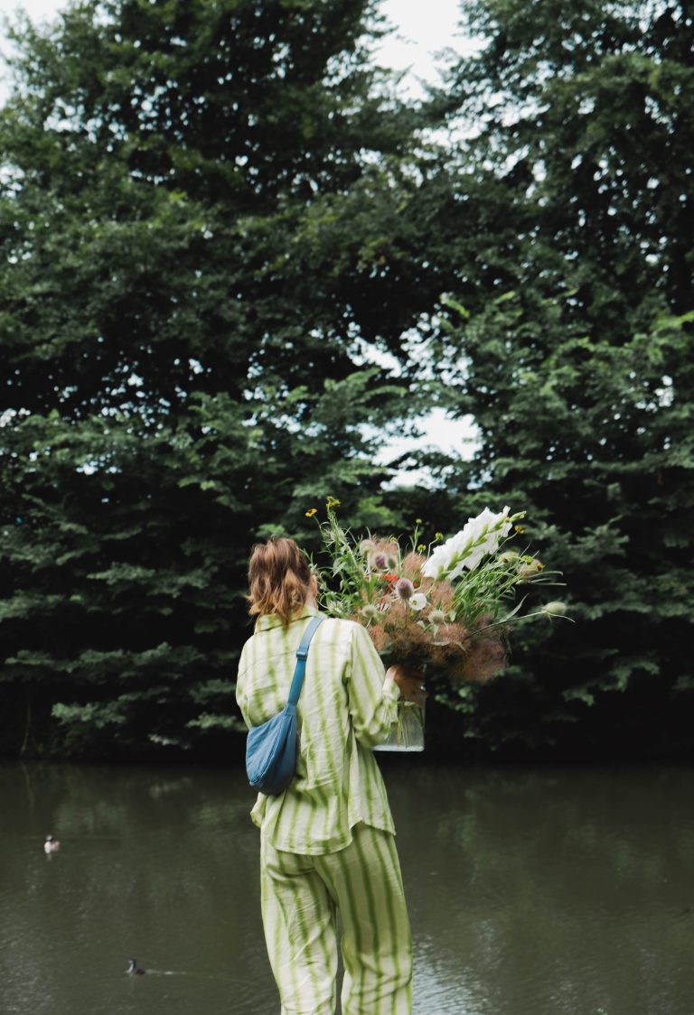 Een persoon in een groen-wit gestreept pak met een blauwe tas draagt een groot veldboeket van Pluktuin Glitter Gladiool, gestyled door Noraly. De vaas met bloemen wordt meegenomen langs het water, met groene bomen en eenden in de achtergrond.