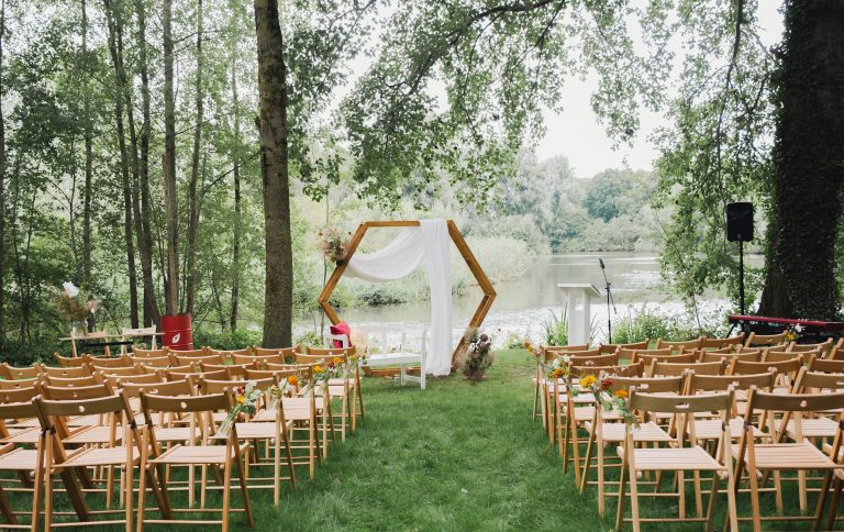 Een sfeervolle trouwceremonie in de natuur, met houten stoelen versierd met bloemen uit Pluktuin Glitter Gladiool. De ceremonieplek is aangekleed door Noraly en heeft een zeshoekige trouwboog met een witte doek en een natuurlijk bloemstuk, uitkijkend over een rustig meer.
