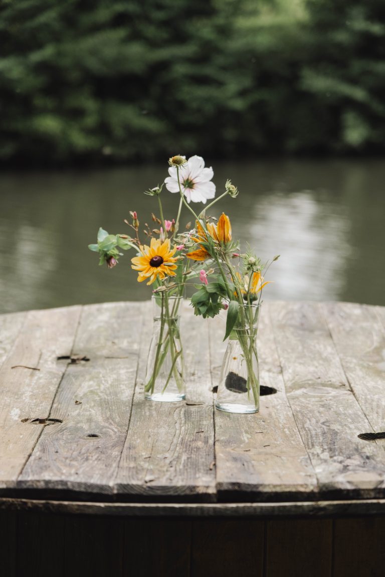 Twee glazen vaasjes met een speelse mix van bloemen uit Pluktuin Glitter Gladiool, waaronder witte cosmos en gele rudbeckia, gestyled door Noraly. De vaasjes staan op een rustieke houten tafel met een waterpartij op de achtergrond.