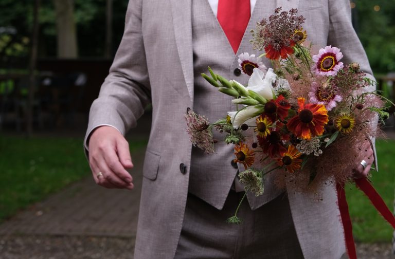 Een bruidegom in een beige pak met rode stropdas houdt een speels, natuurlijk trouwboeket vast, samengesteld door Noraly met bloemen van Pluktuin Glitter Gladiool. Het boeket bevat witte gladiool, rode en oranje rudbeckia en luchtige takken.