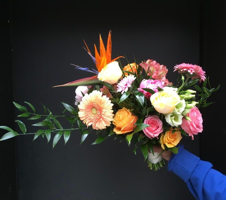 Een kleurrijk handboeket van Oogst met pastel- en felgekleurde bloemen zoals rozen, gerbera’s en een opvallende paradijsvogelbloem, vastgehouden door een persoon in een blauwe trui tegen een donkere achtergrond.