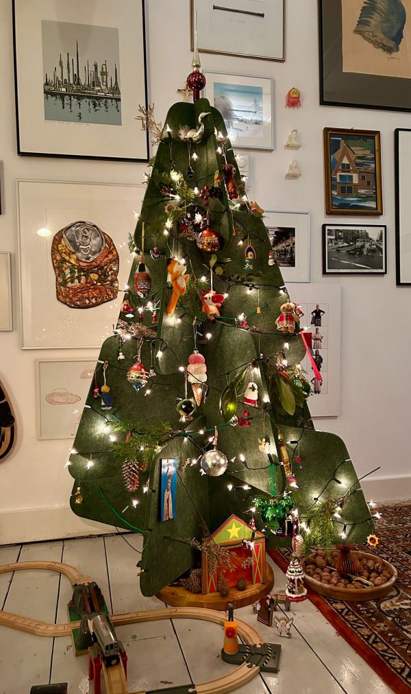 Een groene vilten kerstboom van Jingle Tree staat versierd in een gezellige woonkamer. De boom is gedecoreerd met traditionele kerstballen, kleine figuurtjes en lichtjes die hem een warme sfeer geven. Onder de boom ligt een houten speelgoedtrein en enkele natuurlijke decoraties zoals walnoten.